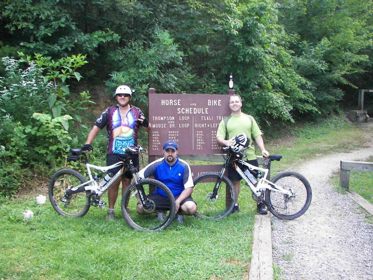 Three people stand near a sign indicating horse and bike schedule for trails, surrounded by lush greenery. Two individuals hold mountain bikes, dressed in athletic gear, while the third person is squatting in front of them. The sign lists trail names and days of the week. Tsali Right Loop mountain bike trail.