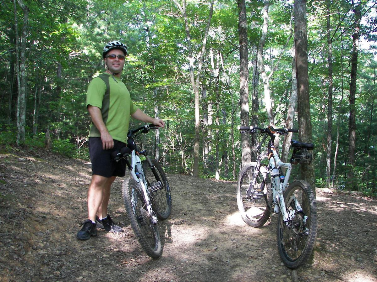 A person stands next to two mountain bikes on a dirt trail surrounded by lush, green trees. The individual is wearing a green shirt, black shorts, and a helmet, smiling as they enjoy the outdoor setting. Tsali Right Loop mountain bike trail.