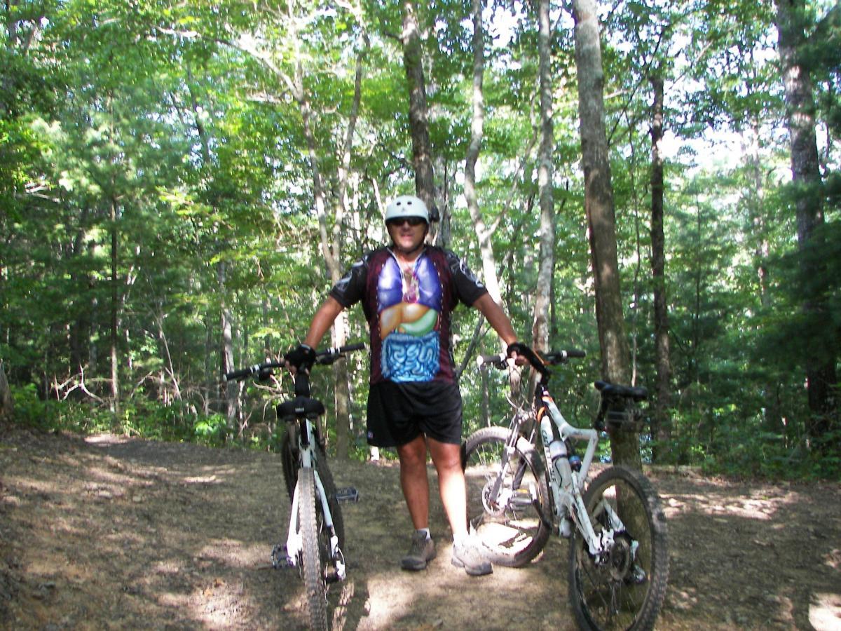 A cyclist dressed in a colorful jersey and helmet stands on a dirt trail surrounded by trees, with two mountain bikes beside him. Sunlight filters through the leaves, creating a vibrant natural setting. Tsali Right Loop mountain bike trail.