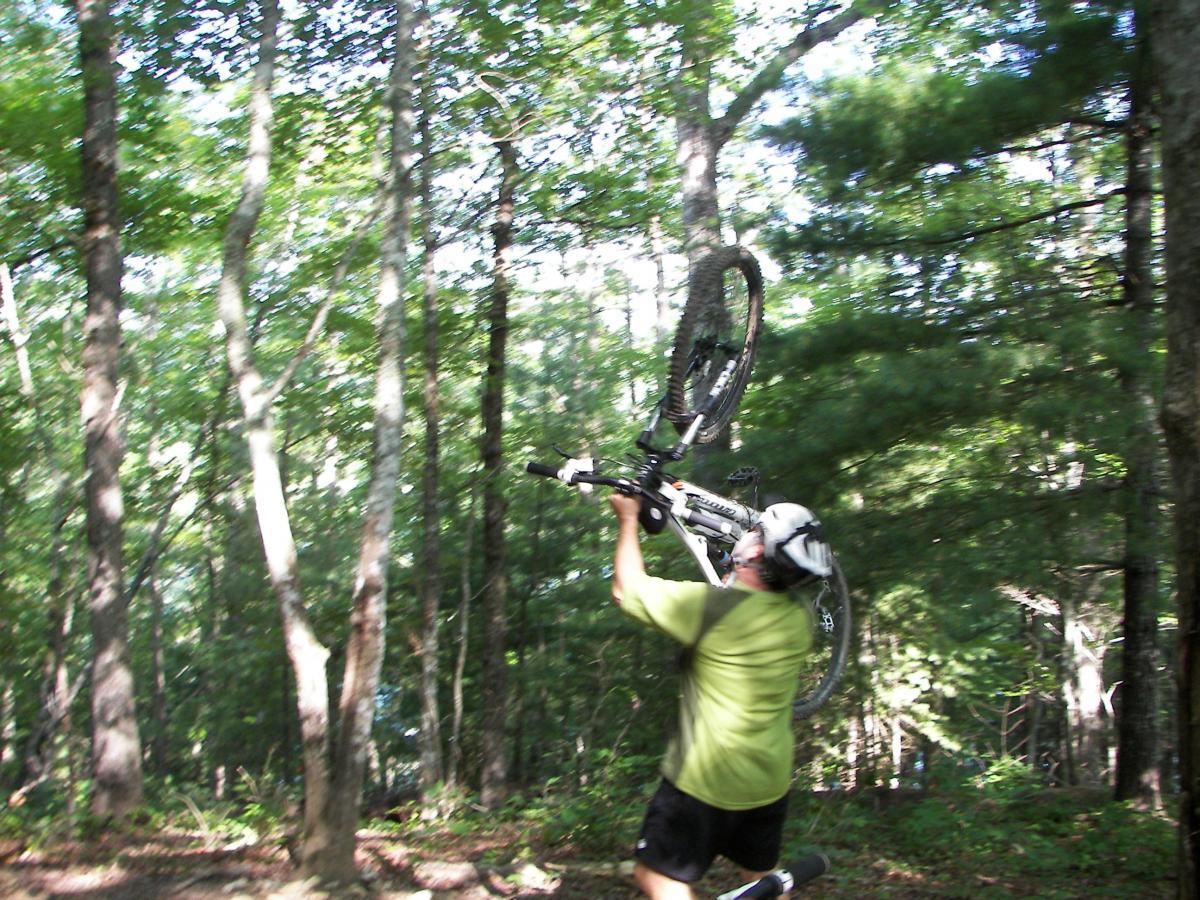 A person wearing a helmet and a green shirt lifts a mountain bike above their head while surrounded by trees in a forested area. The image captures a dynamic moment, highlighting the action of the rider in an outdoor setting. Tsali Right Loop mountain bike trail.