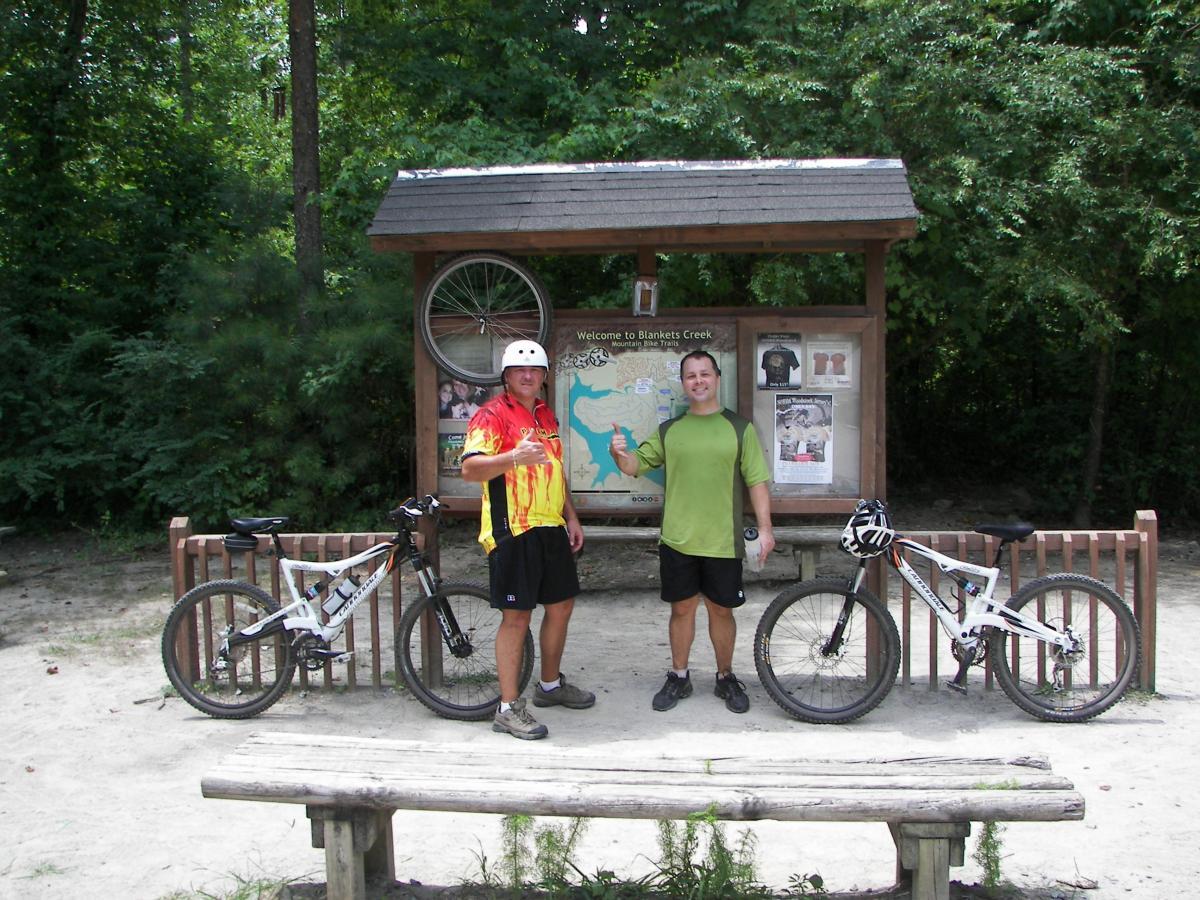 Two cyclists pose in front of a wooden trail sign for Blankets Creek, surrounded by trees. One cyclist, wearing a colorful jersey and helmet, gives a peace sign, while the other, dressed in a green shirt, gives a thumbs up. Their mountain bikes are parked beside them, and a wooden bench is in the foreground. Blankets Creek mountain bike trail.