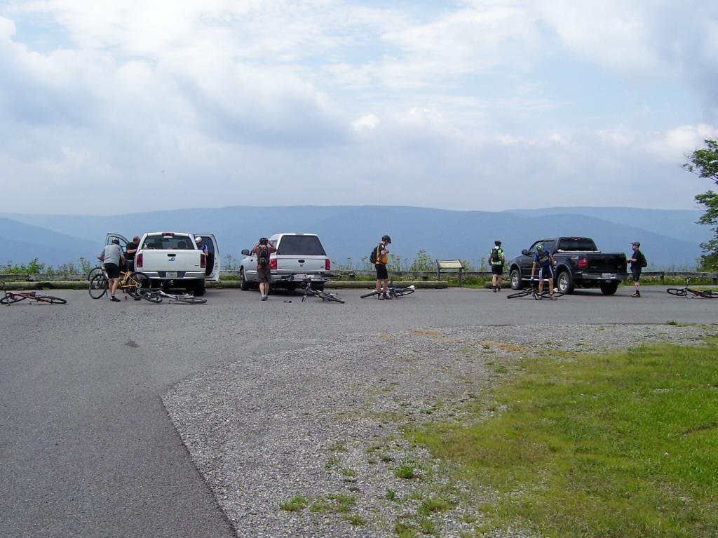 A group of cyclists is parked near several vehicles at a scenic overlook, with their bikes scattered on the ground. The backdrop features rolling mountains under a partly cloudy sky. Some cyclists are retrieving items from the truck beds, while others are mingling or preparing to ride. Gauley-bearpen-boundary-shoals-loop mountain bike trail.