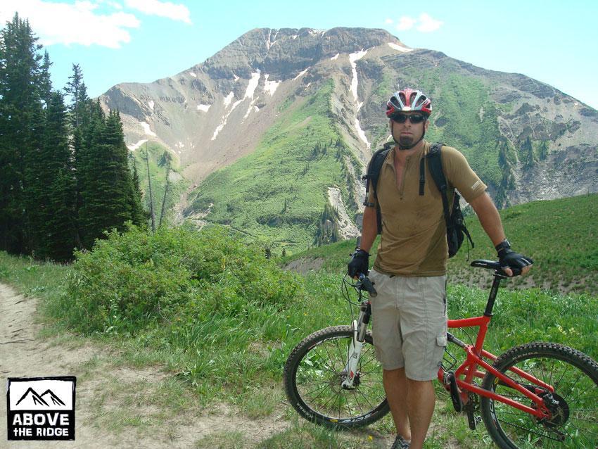A mountain biker stands on a trail with a red mountain bike, wearing a helmet and cycling attire. In the background, a lush green mountain with snow-capped peaks rises against a blue sky. The scene captures the beauty of nature and the excitement of outdoor adventure. The logo "Above the Ridge" is visible in the corner. Trail 401 mountain bike trail.