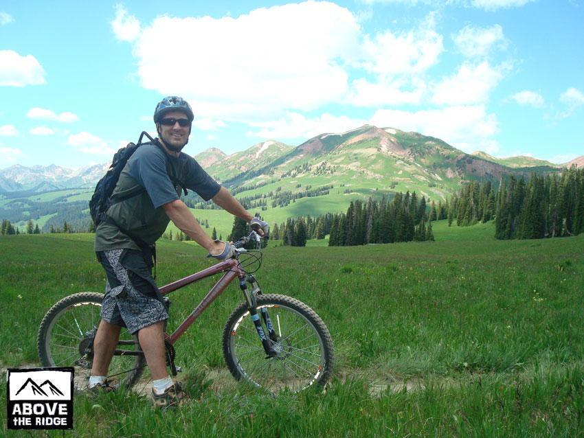 A smiling cyclist stands next to a mountain bike in a lush green meadow, surrounded by scenic mountains and blue skies with fluffy clouds. The cyclist is wearing a helmet and casual outdoor clothing, ready for an adventure in nature. Trail 401 mountain bike trail.