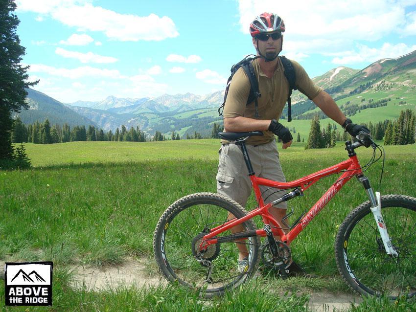A man in a tan shirt and black gloves stands beside a red mountain bike on a grassy trail, surrounded by a scenic landscape of mountains and blue sky with clouds. The area is lush and green, indicating a beautiful outdoor setting for cycling. Trail 401 mountain bike trail.