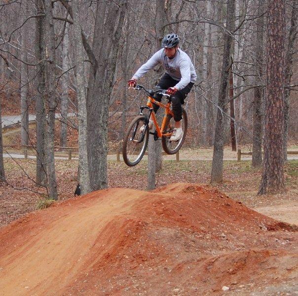 A person performing a jump on a mountain bike over a dirt ramp in a wooded area, with bare trees in the background and a gravel path visible. The cyclist is wearing a helmet and athletic gear. Dirt Jumps mountain bike trail.