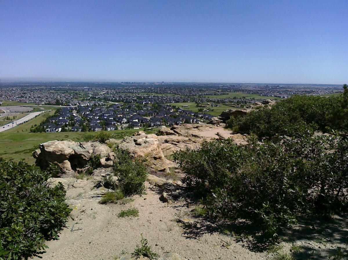 A panoramic view from a rocky outcrop overlooking a suburban landscape, featuring numerous houses with dark roofs, expansive green areas, and a clear blue sky. In the distance, a highway can be seen, along with hints of a city skyline. The foreground is adorned with shrubs and boulders. Wildcat Mountain / Monarch mountain bike trail.