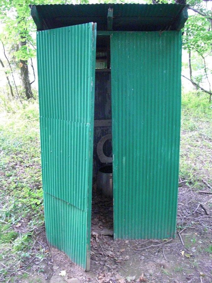 A green, corrugated metal outhouse with two open doors, situated in a wooded area. The interior is partially visible, showing a basic toilet setup. Surrounding the structure is a natural landscape with grass and foliage. Scales Lake Park Trails mountain bike trail.