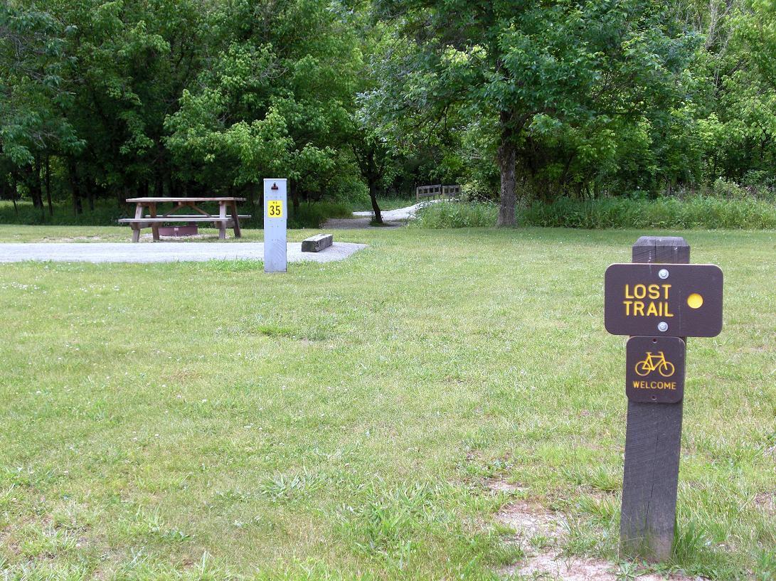 A grassy area featuring a picnic table, a numbered marker labeled "35," and a sign for "Lost Trail" with a bike icon. The scene is surrounded by trees and a pathway leading into the woods. Beech Fork State Park mountain bike trail.