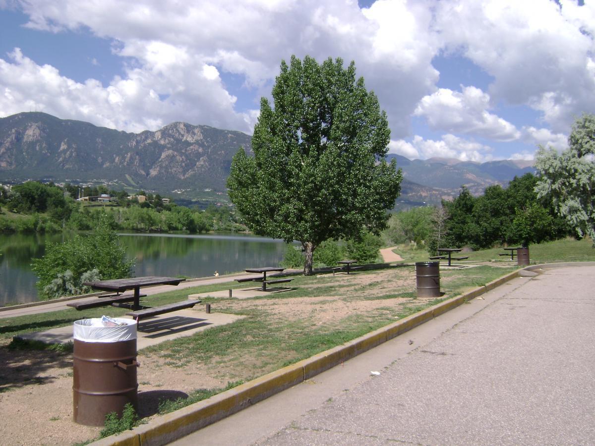 Scenic view of a park by a calm lake, with picnic tables, trash bins, and lush greenery in the foreground. Majestic mountains rise in the background under a partly cloudy sky. Quail Lake mountain bike trail.