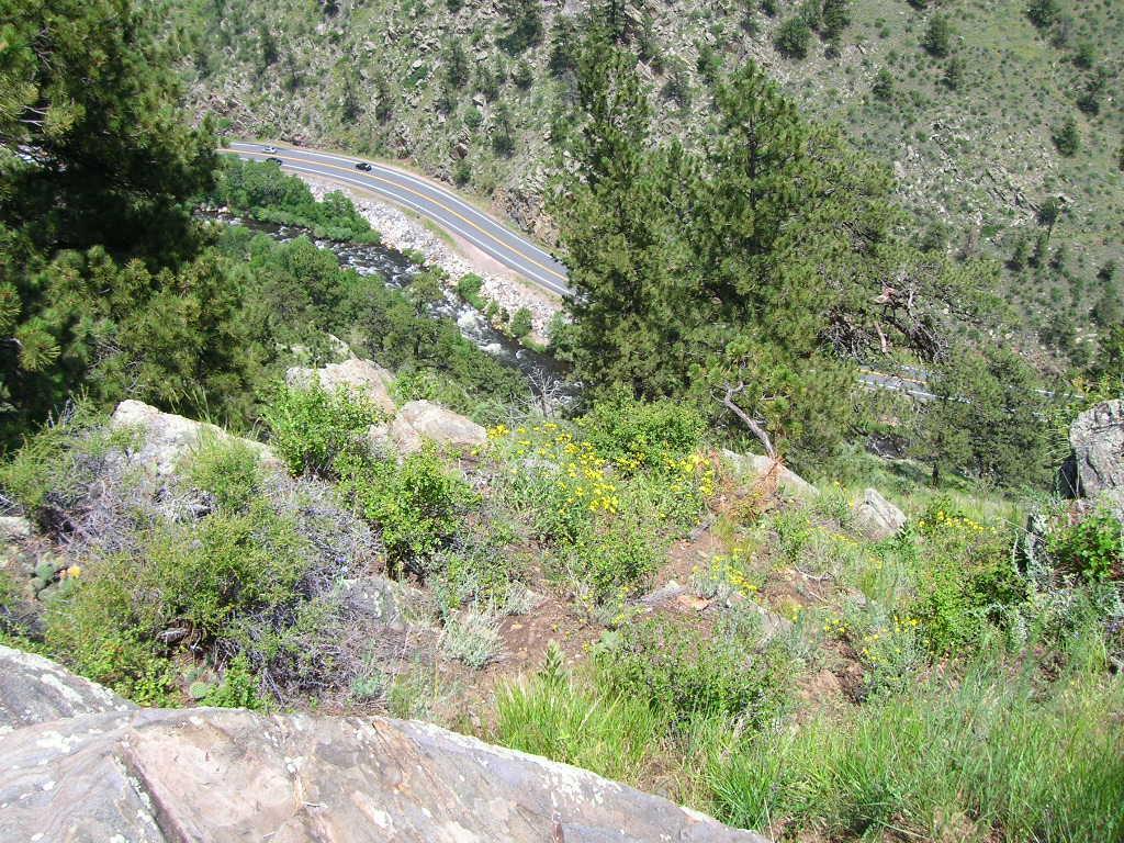 A view from the edge of a rocky cliff overlooking a winding road and a river below, surrounded by greenery, shrubs, and trees on the hillside. Round Mountain Summit Adventure Trail mountain bike trail.