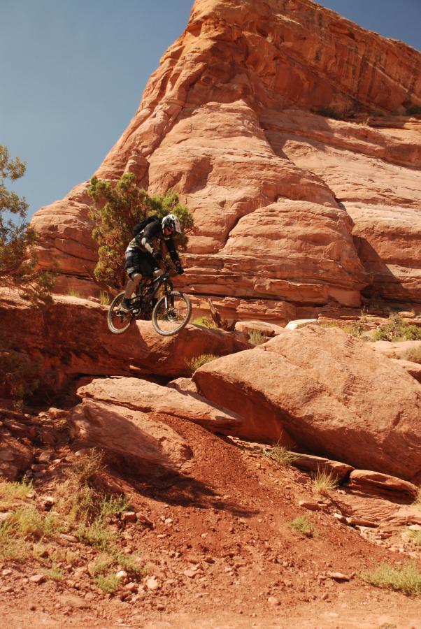 A mountain biker in mid-air jumps over a rocky terrain with red rock formations in the background. The scene captures the thrill of off-road biking in a rugged landscape. Amasa Back Trail mountain bike trail.