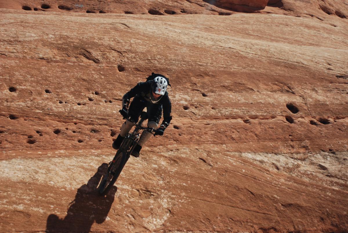 Person riding a mountain bike on a sandy, rocky surface, with a focus on balance and movement. The background features a textured landscape, highlighting the rider's dynamic posture and the rugged terrain. Amasa Back Trail mountain bike trail.