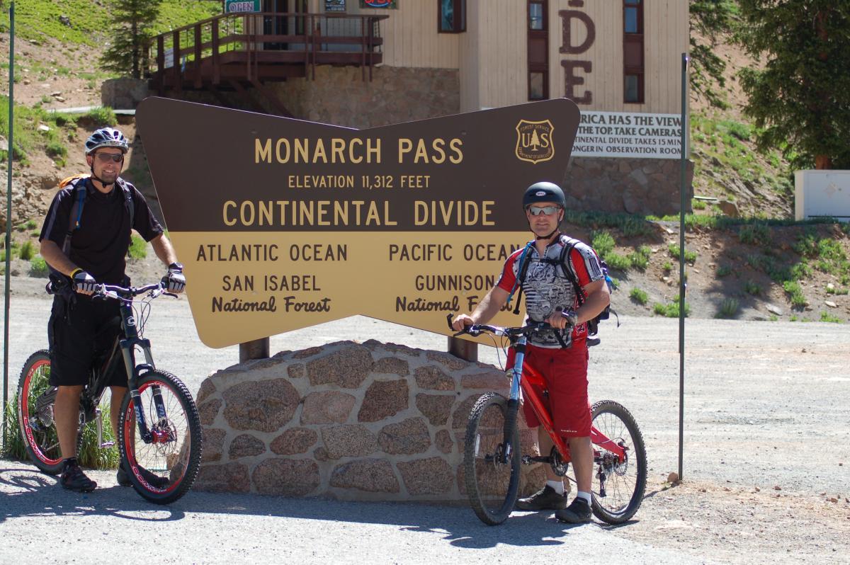 Two mountain bikers pose next to a large sign at Monarch Pass, which marks the Continental Divide at an elevation of 11,312 feet. The sign displays information about the Atlantic and Pacific Oceans and the nearby San Isabel and Gunnison National Forests. In the background, a building and greenery are visible, indicating a mountainous outdoor setting. Monarch Crest Trail mountain bike trail.