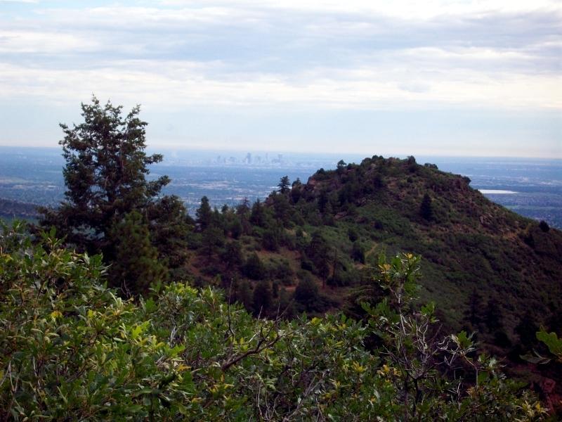 A scenic view from a mountain lookout, featuring a green hillside with trees in the foreground and a distant city skyline visible on the horizon under a cloudy sky. Deer Creek Canyon mountain bike trail.