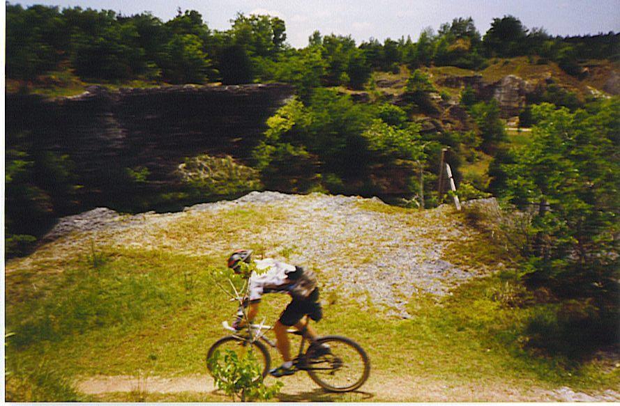 A person riding a mountain bike on a gravel path surrounded by greenery and rocky terrain. In the background, there are cliffs and trees under a clear sky. Haile's Trails mountain bike trail.