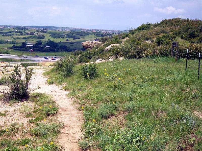 A scenic view of a grassy hillside with wildflowers leading down to a road. In the background, rolling hills and a blue sky with scattered clouds are visible. A few parked cars are on the road, while bushes and rocky outcrops are present on the hillside. Glendale Open Space Trail mountain bike trail.