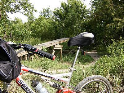 A close-up view of a bicycle's seat and handlebars in a natural setting, with a wooden bridge visible in the background surrounded by greenery. Bonnet Carre Spillway Trail mountain bike trail.