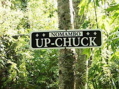 A sign reading "NOMAMBO UP-CHUCK," mounted on a tree, surrounded by lush greenery in a forested area. Bonnet Carre Spillway Trail mountain bike trail.