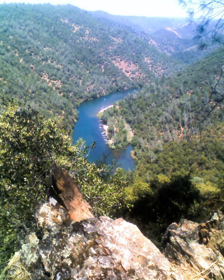 A panoramic view of a winding river flowing through a forested valley, surrounded by lush green hills and rocky outcrops under a clear blue sky. Clementine / Forresthill Connector Trail mountain bike trail.