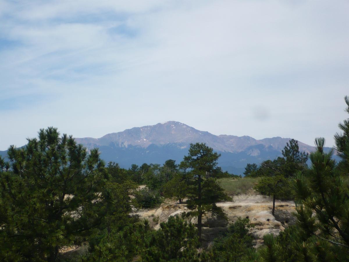 A scenic view of distant mountains, partially covered in snow, rising above a foreground of lush green trees and rocky terrain under a partly cloudy sky. Palmer Park mountain bike trail.