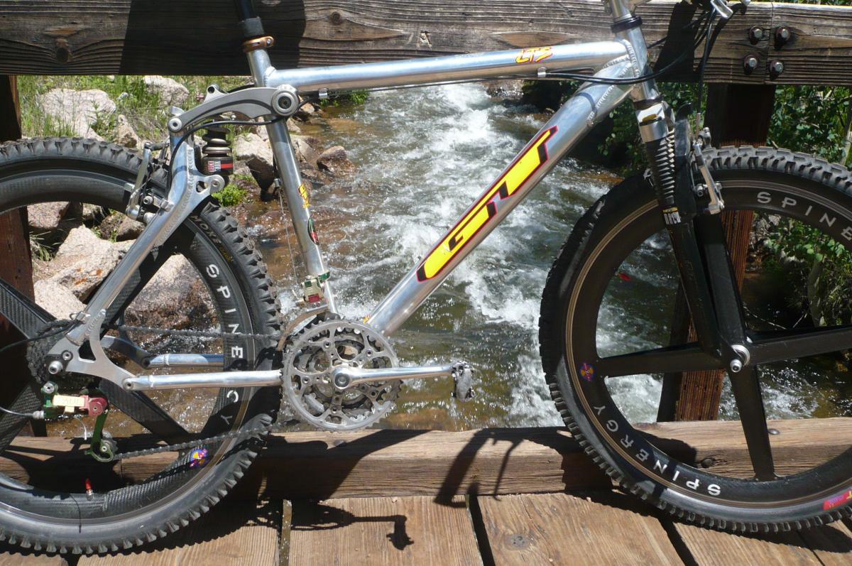 Mountain bike leaning on a wooden bridge, with a flowing stream in the background. The bike features a shiny aluminum frame, large knobby tires, and visible components like the chain and suspension system, set against a natural outdoor setting. Rampart Reservoir mountain bike trail.