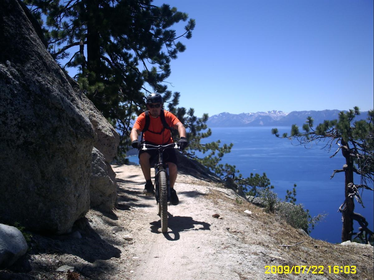 A mountain biker in an orange shirt and helmet rides along a narrow, scenic trail bordered by large rocks and trees, with a panoramic view of a blue lake and distant mountains in the background under a clear sky. Flume Trail mountain bike trail.