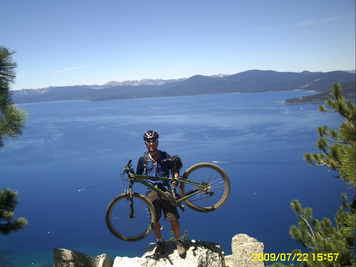 A mountain biker standing on a rocky ledge overlooking a clear blue lake and mountains in the background. The biker is holding a bicycle and wearing a helmet and sunglasses, with trees visible nearby. The scene is bright and sunny, showcasing a beautiful outdoor landscape. Flume Trail mountain bike trail.