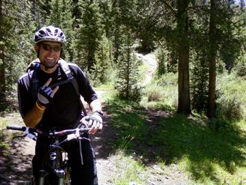A mountain biker wearing a helmet and gloves smiles while leaning on his bicycle in a wooded area. Behind him, a winding trail can be seen through the trees. The scene is sunny with green grass and foliage surrounding the path. Monarch Crest Trail mountain bike trail.