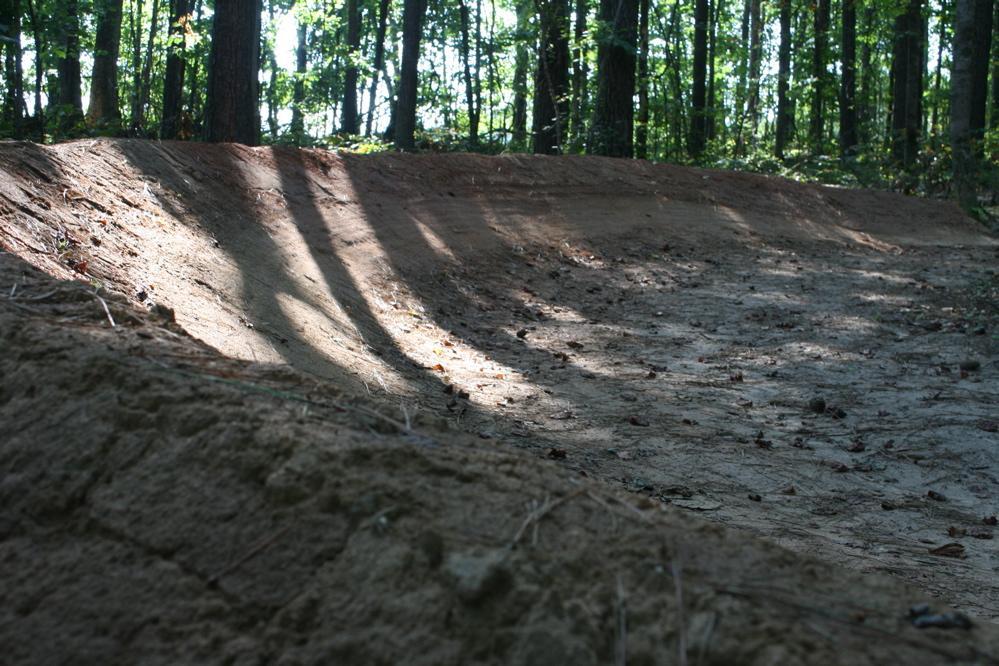 A sunlit dirt bike track winding through a wooded area, with long shadows cast by trees. The ground is uneven and sandy, featuring scattered leaves and small rocks, and the trail slopes gently upwards. Harbins Park mountain bike trail.