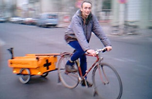 A person riding a bicycle with a large orange trailer attached, moving along a street. The cyclist is wearing a gray jacket and blue pants, with a focused expression. The background shows blurred parked cars and buildings, indicating motion.
