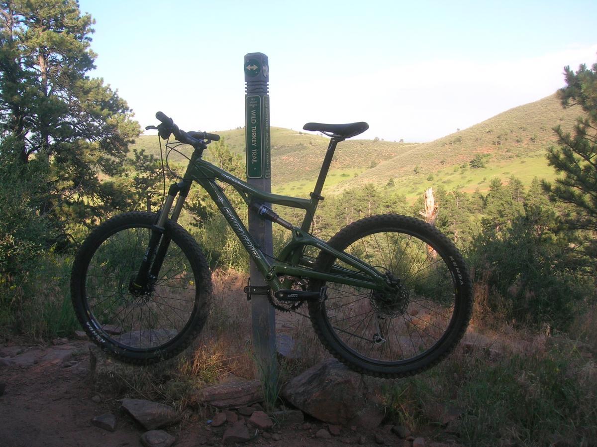 A green mountain bike leaning against a trail sign that reads "Wild Turkey Trail," set against a backdrop of rolling hills and trees in a natural landscape. Heil Valley Ranch mountain bike trail.