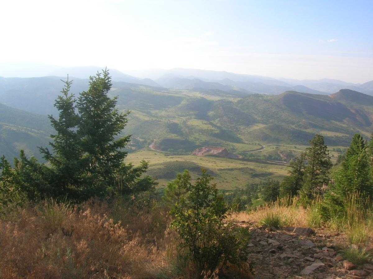 A scenic mountain landscape featuring rolling hills and valleys, with lush greenery in the foreground. Pine trees and shrubs can be seen among rocky terrain, while distant mountains fade into a soft, blue sky. The sunlight casts a gentle glow across the scene, suggesting a clear day. Heil Valley Ranch mountain bike trail.