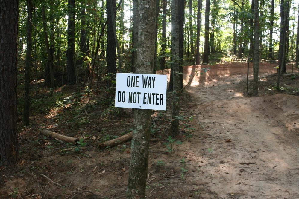 A sign on a tree in a wooded area that reads "ONE WAY DO NOT ENTER." The path in the background is partially visible, indicating a trail that curves away from the sign. The surrounding forest features tall trees and scattered foliage. Harbins Park mountain bike trail.