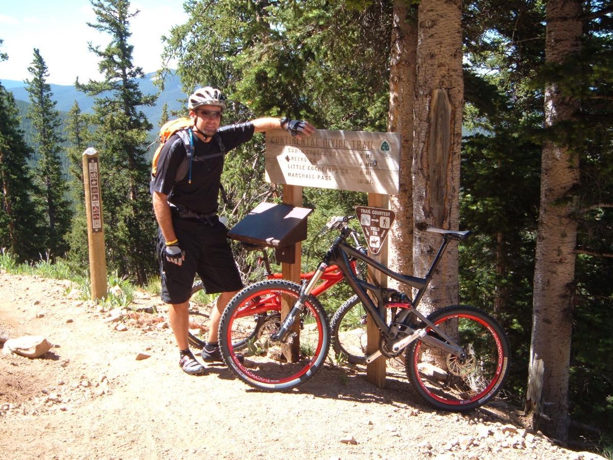 A mountain biker in a black jersey and helmet, standing next to a bike with red wheels, beside a trail sign indicating the Continental Divide Trail. The background features a scenic view with trees and mountains under a clear blue sky. Monarch Crest Trail mountain bike trail.