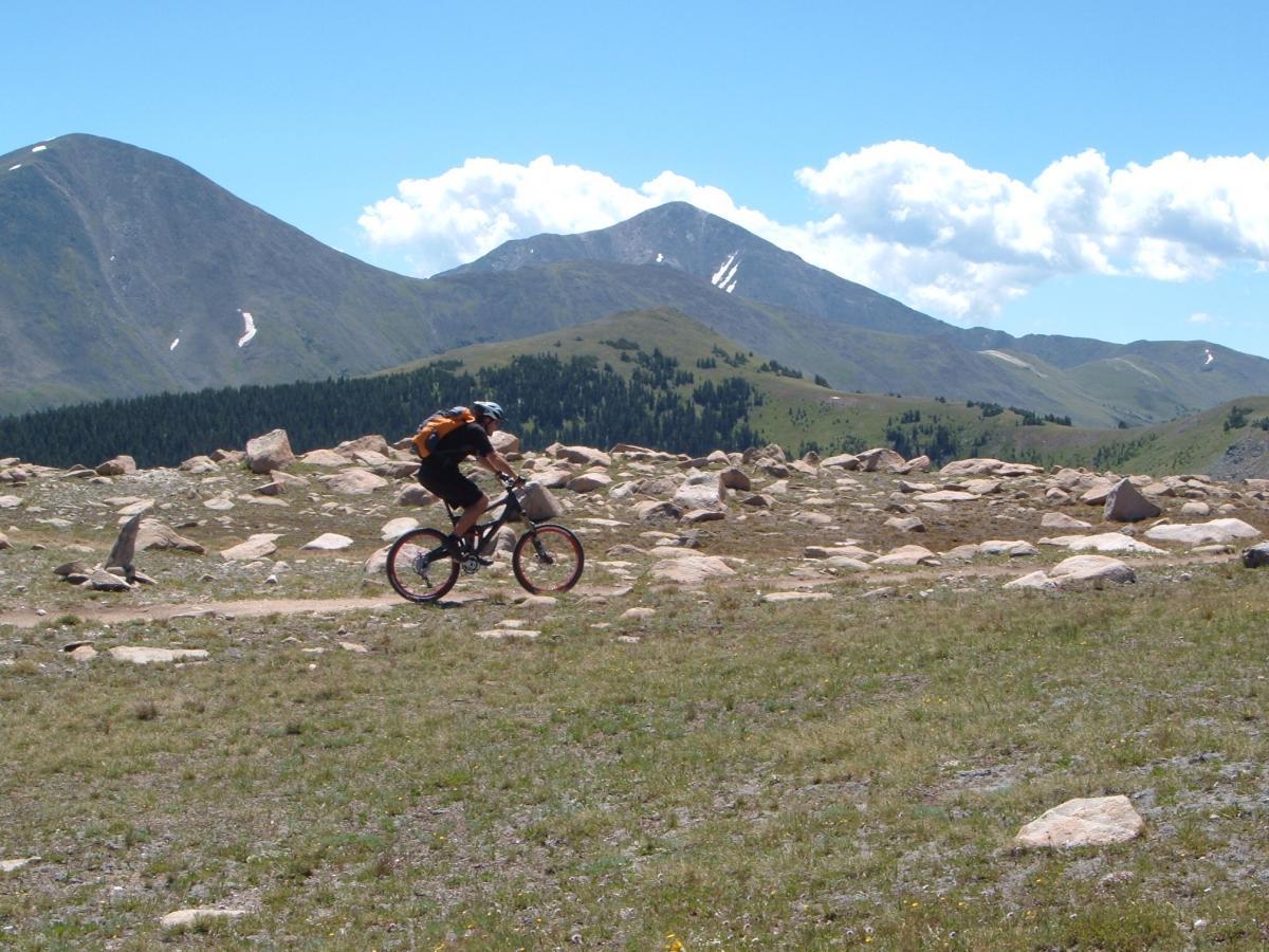 A mountain biker rides along a rugged trail surrounded by rocky terrain and rolling green hills under a clear blue sky with scattered clouds and distant mountains. Monarch Crest Trail mountain bike trail.