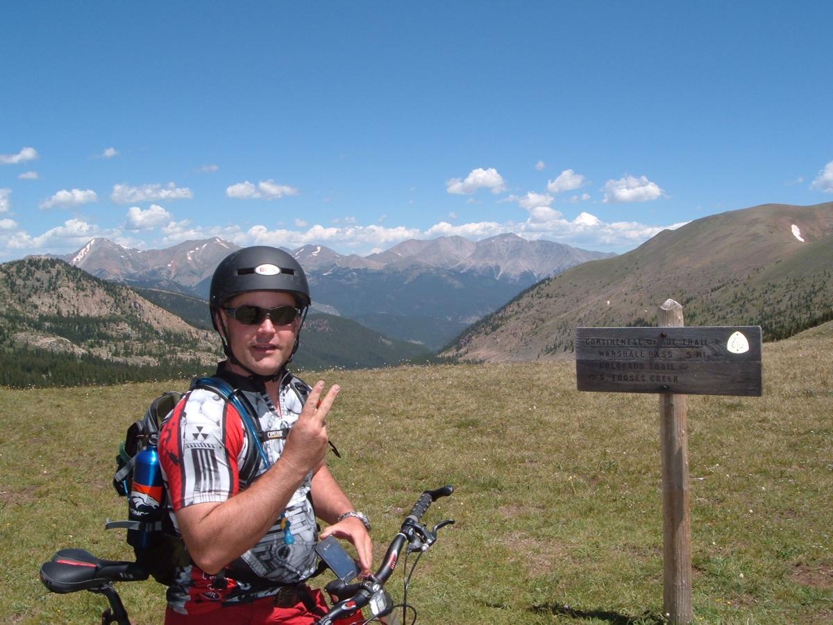 A person wearing a helmet and sunglasses poses with a bicycle, giving a peace sign. In the background, snow-capped mountains and a clear blue sky create a scenic view. A wooden sign nearby indicates the location of the Continental Divide Trail at Marshall Pass, Colorado. Monarch Crest Trail mountain bike trail.