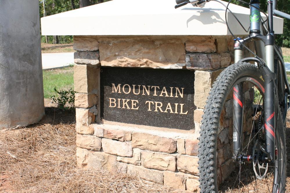 Stone monument with a plaque reading "Mountain Bike Trail," alongside the front wheel of a mountain bike. The background features a dirt path and lush greenery. Harbins Park mountain bike trail.