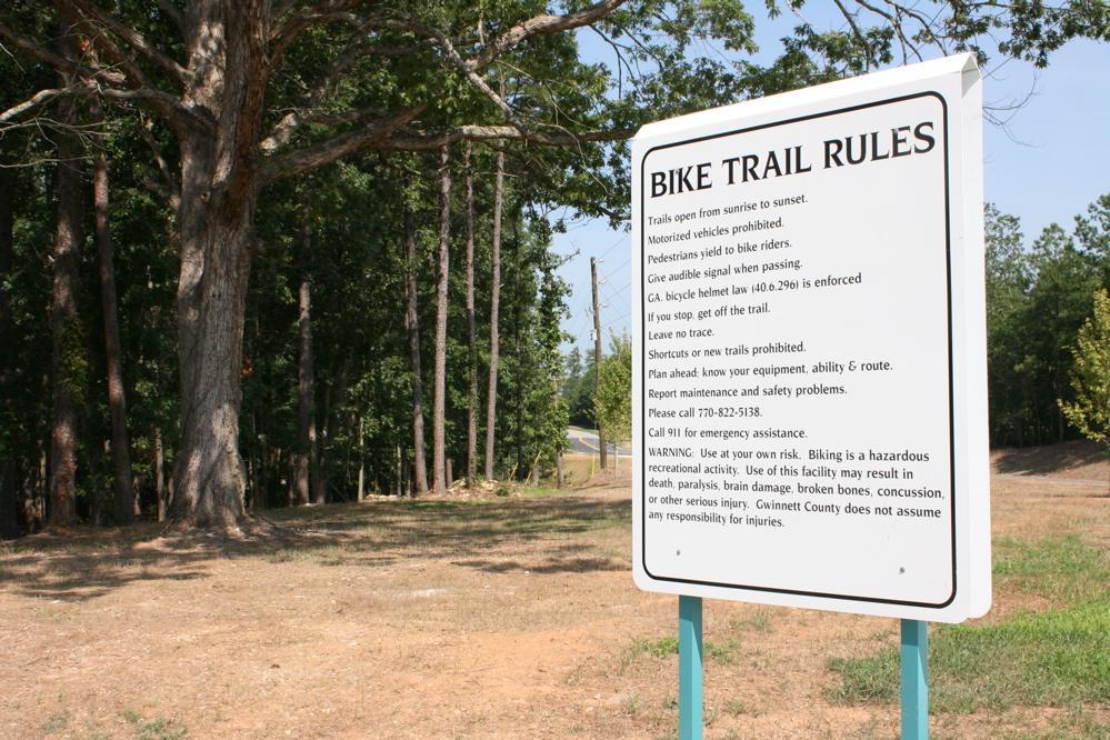 A sign listing the bike trail rules, including instructions for trail usage, safety warnings, and contact information for emergencies, set against a background of trees and open land. The sign emphasizes the need for caution and respect for the trail environment. Harbins Park mountain bike trail.