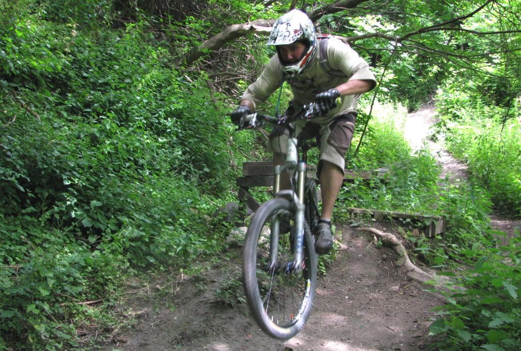 A mountain biker performing a jump on a dirt trail surrounded by lush greenery. The rider is wearing a helmet and protective gear, showcasing an action-packed moment in outdoor biking. Don Valley mountain bike trail.
