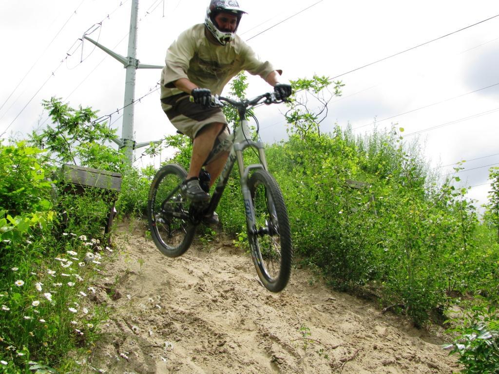 A mountain biker performing a jump on a dirt trail surrounded by greenery and wildflowers, with power lines in the background. The rider is wearing a helmet and protective gear, and is captured mid-air above a sandy slope. Don Valley mountain bike trail.