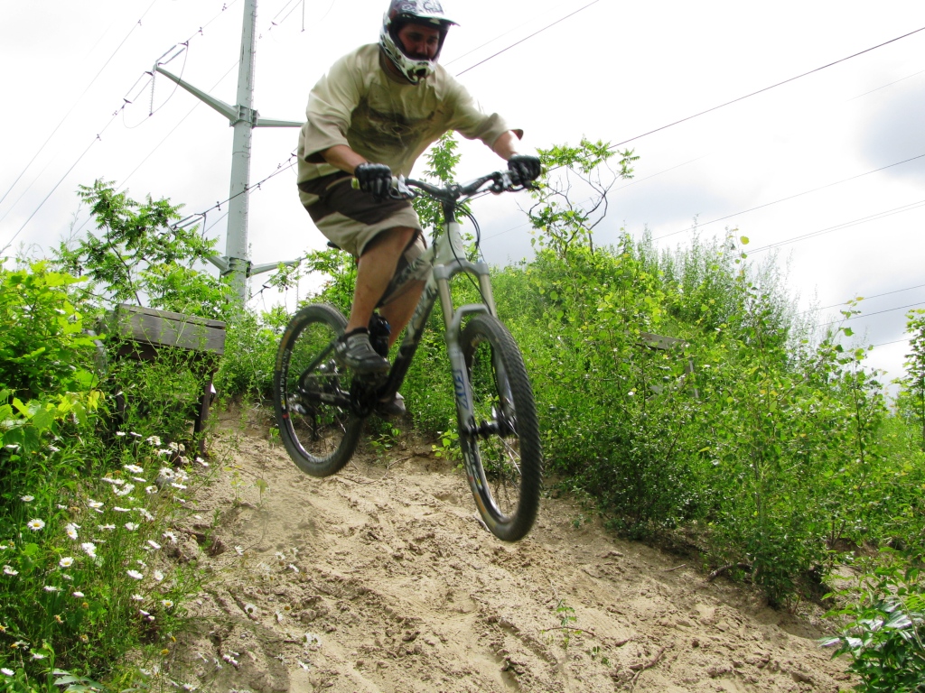 A mountain biker in mid-air jumps off a sandy trail surrounded by greenery, with power lines in the background. The cyclist is wearing a helmet and protective gear, showcasing an action-packed moment in outdoor biking.