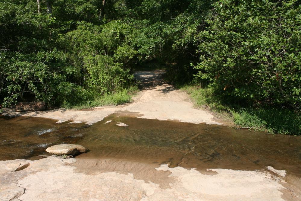A serene natural landscape featuring a shallow stream with clear water, flowing over a sandy and rocky riverbed. Lush green foliage and trees line the banks of the stream, creating a peaceful and inviting atmosphere. A narrow path leads away from the water, hinting at exploration opportunities in the surrounding nature. Harbins Park mountain bike trail.