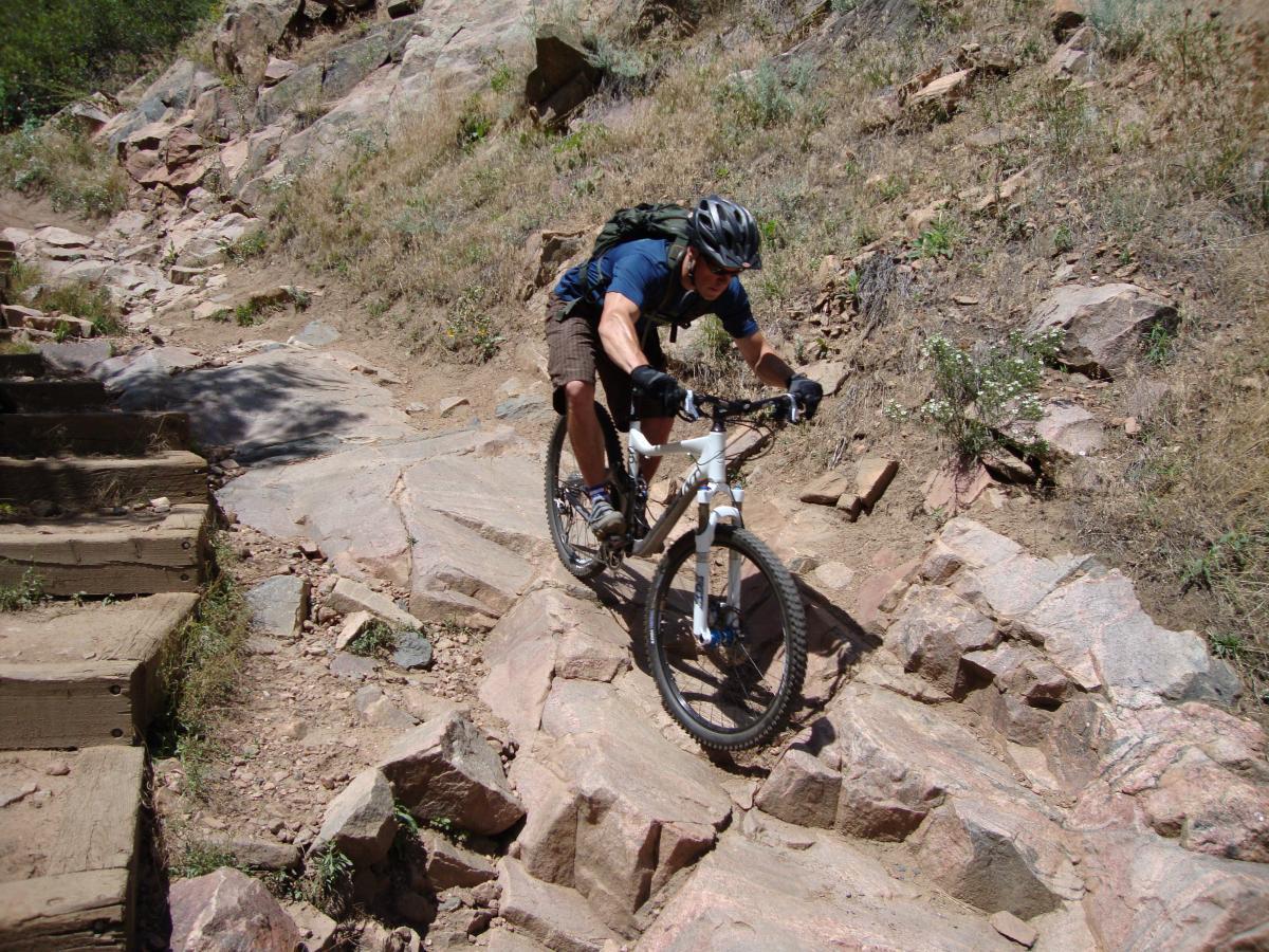 A mountain biker navigating a rocky trail, wearing a helmet and a backpack, with steep terrain and natural vegetation in the background. Deer Creek Canyon mountain bike trail.