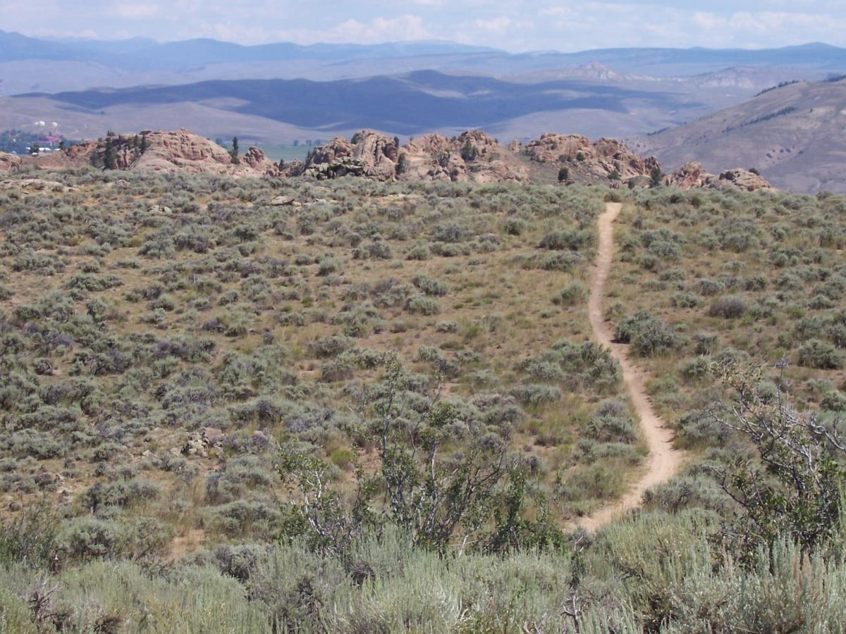 A winding dirt path leading through a vast expanse of sagebrush and grasslands, with rocky outcrops visible in the background against rolling hills under a clear blue sky. Hartman Rocks mountain bike trail.