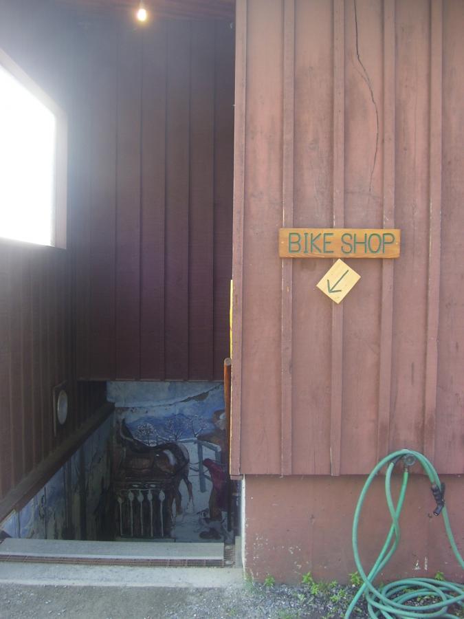 A wooden building exterior featuring a sign that reads "BIKE SHOP" with a downward arrow, indicating the entrance to a bike shop located at the bottom of a staircase. The entrance is partially visible, showcasing a colorful mural on the wall nearby. A green garden hose is coiled on the ground beside the entrance.