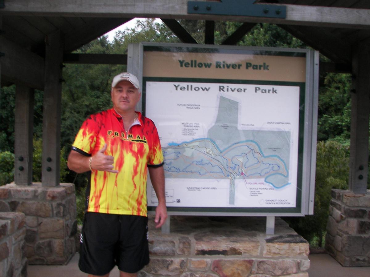 A man in a colorful flame-patterned cycling jersey stands next to a park map at Yellow River Park, giving a thumbs-up. The map displays trails and features of the park, set against a backdrop of trees. Yellow River mountain bike trail.