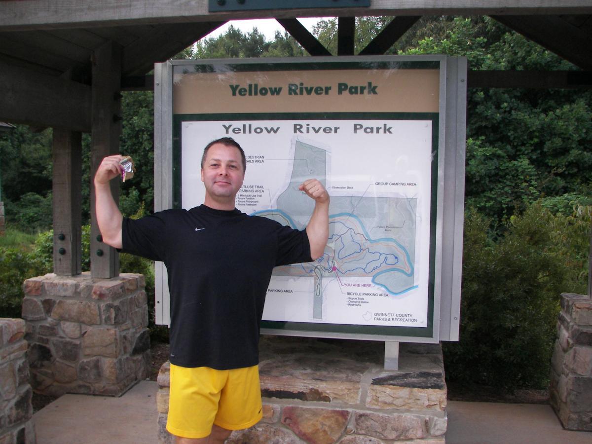 A man in a black t-shirt and yellow shorts stands in front of a park map for Yellow River Park, flexing his muscles with a smile. The background features greenery and a stone structure. Yellow River mountain bike trail.