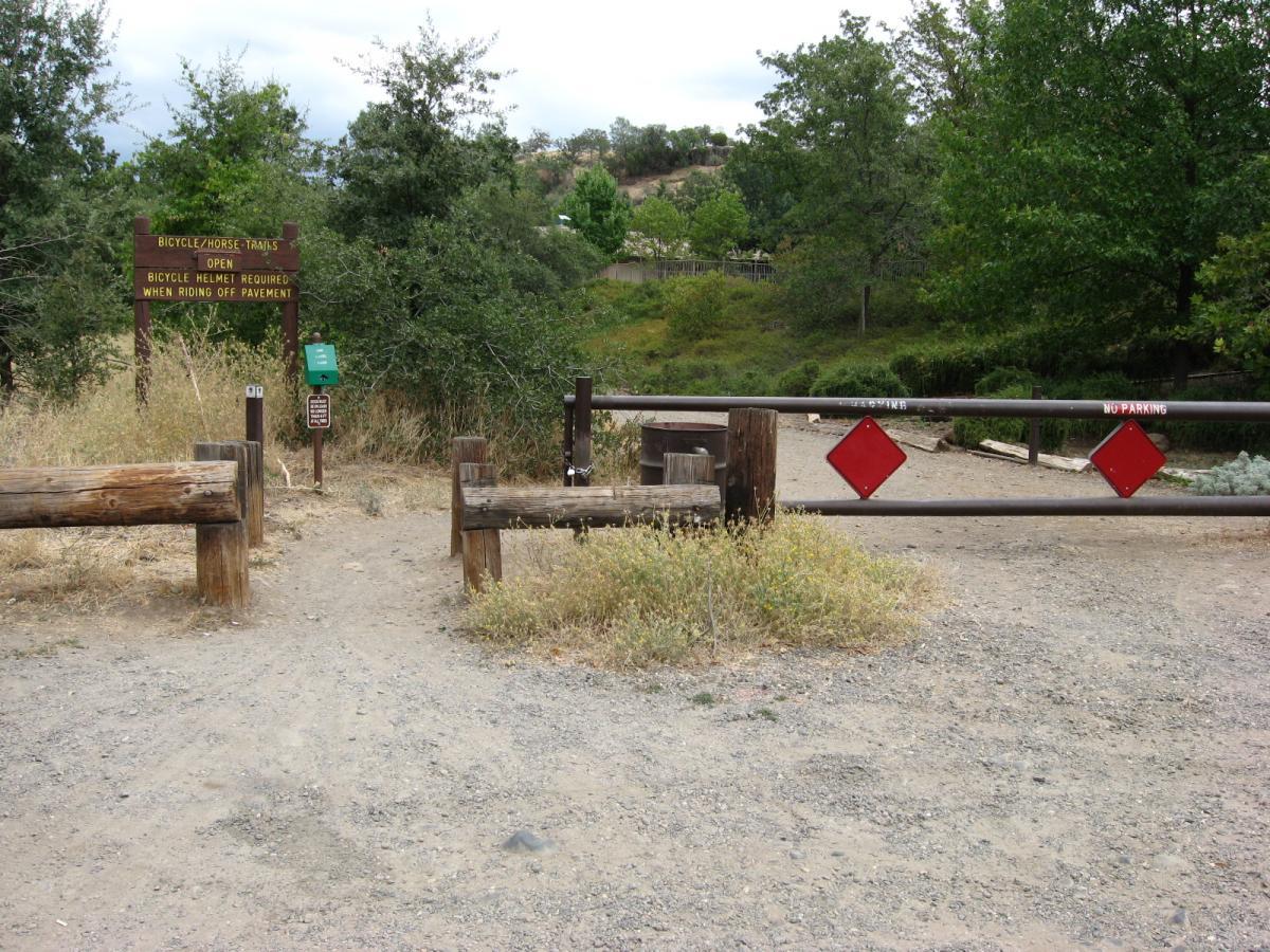 Alt text: Entrance to a trail marked for bicycles and horses, featuring a wooden sign indicating trail rules, a green information box, and a locked gate with red diamond warning signs. Surrounding area includes grass and trees under a cloudy sky. Upper Bidwell Park mountain bike trail.