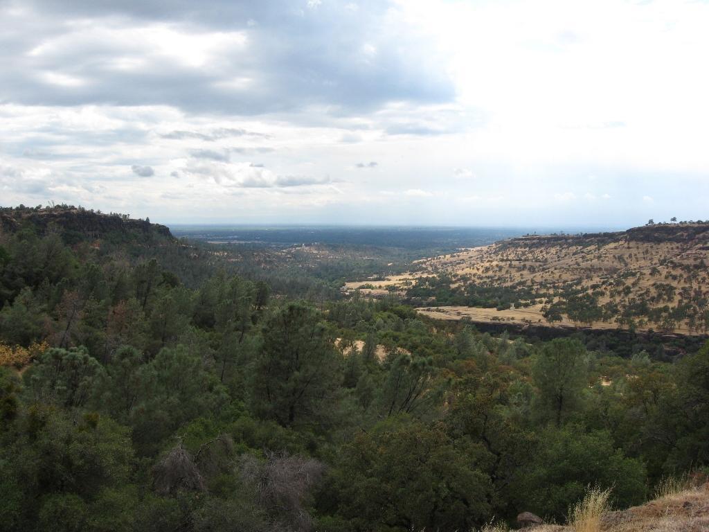 A panoramic view of a mountainous landscape featuring rolling hills, dense patches of green trees, and open areas of dry grassland under a cloudy sky. The scene captures the natural beauty of the outdoors. Upper Bidwell Park mountain bike trail.
